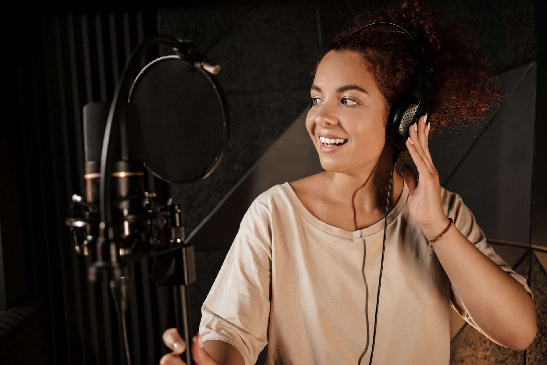 Eine Person mit lockigem Haar und Kopfhörern steht vor einem Mikrofon und scheint in einem Tonstudio zu singen oder zu sprechen.